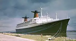 Photographie en couleurs d’un paquebot noir et blanc avec des cheminées rouges et blanches amarrés à un quai en herbe sous un ciel orageux.