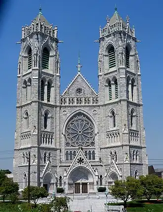 Photographie couleur d’une grande église, grise, assez massive, flanquée de deux tours jumelles à base carrée tournées d’un huitième de tour par rapport à la façade (offrant donc à la vue leurs arrêtes).
