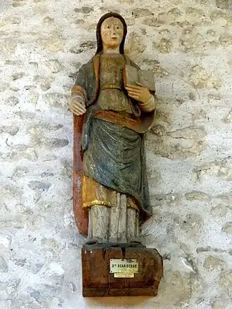 statue disposée en hauteur sur un socle au mur ; elle représente une femme debout, aux cheveux sombres, vêtue d’une robe drapée blanche, bleue et rouge, et portant un livre ouvert dans la main gauche