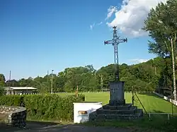 Monument avec une croix en fer forgé. Au centre est représenté le Sacré-Coeur de Jésus, au sommet est placé un coq. En arrière plan, le stade de football
