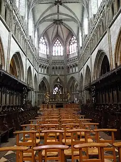 Photographie d'un choeur d'église avec le maître-autel, des stalles et le centre occupé par des chaises.