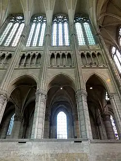Trois piliers sont visibles reliés par un mur de pierre. Au-dessus, le triforium et quatre fenêtres hautes de verre blanc.