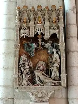 Sculpture représentant, dans un baldaquin sculpté et en partie doré, six personnages surmontés par deux petits anges. Parmi ces personnages, deux sont debout tandis que le troisième est au sol, soutenu par le quatrième personnage, une femme portant une robe bleue et rouge. Les deux derniers personnages, plus petits, sont agenouillés et prient. La sculpture est dans un état dégradé.