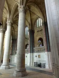 Au premier plan, des piliers, dont certains sont peints liés par des tirants de bois. En arrière-plan, un mur massif orné de bas-reliefs.