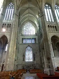 Mur très haut percé de plusieurs grandes verrières. En bas de l'image, bancs en bois et statues de pierre. En haut de l'image, grande rosace illuminée par le soleil.