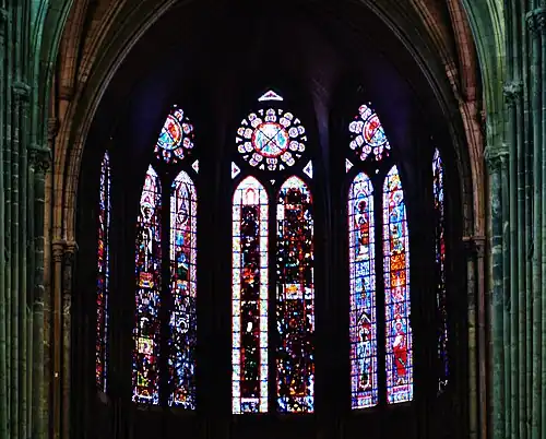 Vue depuis la nef des verrières des fenêtres hautes du chœur. L'image est sombre et de mauvaise qualité. Sont cependant visibles, sur la seconde fenêtre en partant de la droite, quatre hommes portant des auréoles. De part et d'autre des verrières, des piliers de la nef sont visibles.
