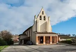 Photo de l'église Saint-Pierre : vue de la porte d'entrée, protégée par un porche avec des colonnes en briques et un toit en tuile, sous le clocher-mur qui abrite trois cloches. Une croix surmonte le clocher-mur.