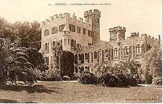 Château néo-médiéval avec tours crénelées, échauguette et palmiers ; nombreuses fenêtres en façade avec volets ouverts cintrés.