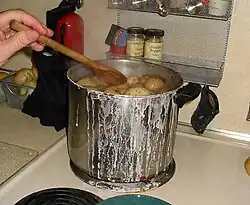 À l’avant-plan, sur la plaque électrique d’un fourneau domestique blanc, une grosse marmite en acier inoxydable et poignées latérales noires est pleine de pommes de terre cuisant dans l’eau qui a débordé et laissé de nombreuses traces blanches sur le corps du récipient et sur la plaque ; on aperçoit la main qui vient de saler l’eau avec une cuiller en bois ; juste derrière le fourneau, suspendue au mur, une petite étagère métallique grillagée contient quelques pots d’épices et supporte deux maniques ; près d’elle, sur le plan de travail, un extincteur rouge et noir, un ravier métallique contenant de gros ognons.