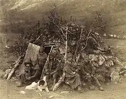 Vue d'un groupe de personnes devant une hutte constituée de pierres et de branches d'abres