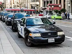 Voitures de patrouilles du SFPD garé au bord d'un trottoir à Sain Francisco. La première est une Ford au capot sombre et aux portières blanches marquées du badge SFPD. Elle est équipé d'avertisseurs lumineux bleus, blancs et rouges sur le toit et de projecteurs au-dessus des rétroviseurs.