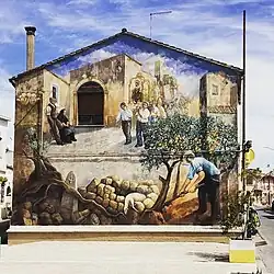 Photographie d'un mur de maison avec sommet triangulaire surmonté sur la gauche d'une haute cheminée longiligne, avec sur toute la surface une peinture représentant en bas à gauche un cultivateur avec béret, au centre un mouton affaissé sur un tas de pierres, à gauche en vrac une stèle, une tête de statue et diverses pièces archéologiques, le tout entremêlé dans une racine menant à un arbre fruitier ; en arrière plan dans un décor villageois, à droite une procession avec des fidèles portant une statue mariale ; sur la gauche un homme et une femme spectateurs, lui appuyé dos au mur, elle assise sur un siège.