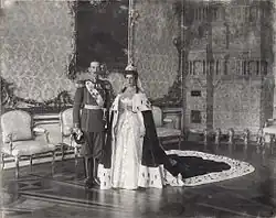 Cliché noir et blanc dans grand salon. Jeune homme : uniforme militaire. Jeune femme en mariée et long manteau impérial.