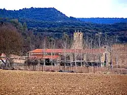 Photographie d'un clocher et de bâtiments agricoles dans un champ ; en arrière-plan, un paysage boisé et vallonné.
