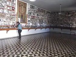 Photographie en couleurs. Intérieur d'une salle, dallée avec des triangles noir et blanc ; sur les murs, de nombreux ex-voto sont accrochés, que consulte une femme.