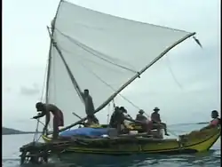 Photographie en couleur d'une pirogue à balancier sur laquelle se trouvent six hommes qui manœuvrent l'embarcation