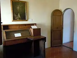 Cellule monacale avec bureau surplombé d'un cadre pieux et petite porte ouverte à droite