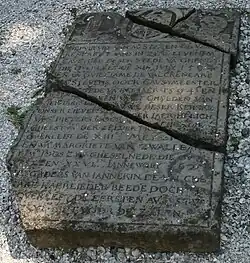 Photographie en couleurs d'une pierre tombale morcelée et gravée d'incriptions