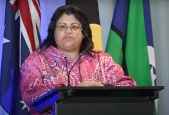 Une femme brune parle à une tribune, avec plusieurs drapeaux derrière elle.