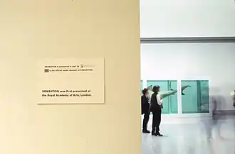 Exposition d'un requin dans une eau verte, un mur devant avec une affiche indiquant que l’œuvre, Sensation des Young British Artists, a d'abord été exposée à l'Académie royale des arts de Londres