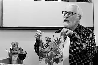 Serge Le Tendre présentant la couverture du tome 1 de l'intégrale de la série, représentant les personnages de Bragon et Pélisse. Un buste en résine de Bragon se trouve à ses côtés.