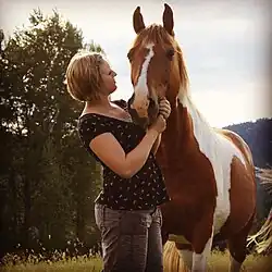Photographie en couleur d'un cheval de couleur rousse et blanche et d'une femme qui lui touche la tête.