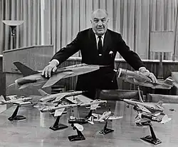 photo noir et blanc montrant un homme assez âgé, en costume, entouré de maquettes d'avions militaires.