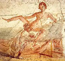 Homme habillé accroupi au bord d'un lit prodiguant un cunnilingus à une femme nue assise sur ce meuble, les jambes écartées