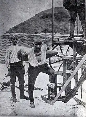 Photographie prise depuis un navire montrant une forteresse en ruine surplombant une pente escarpée menant à une étroite plage.