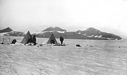 Photo en noir et blanc de deux tentes pyramidales sur un glacier devant un massif montagneux peu élevé.