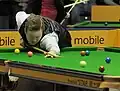 Homme effectuant un tir lointain. La bille blanche a impacté la bille rouge, qui semble se diriger vers la poche du bas, à gauche. À l'arrière plan, on voit un spot publicitaire jaune portant l'inscription « mobile ». On voit aussi une silhouette de dos tenant une queue de billard et un reposoir. Il s'agit probablement d'un homme.