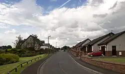 Photo d'une rue résidentielle bordée de maisons individuelles avec des jardins bien entretenus. Un ciel partiellement nuageux est visible au-dessus, et une voiture rouge est garée devant l'une des maisons sur la droite.