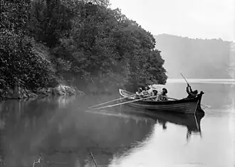 Photographie ancienne en noir et blanc d'une barque quittant un rivage dont les arbres surplombent directement la mer.