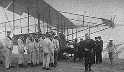 Photographie noir et blanc d'un avion, au sol, avec des matelots autour et un homme, en manteau sombre, qui marche vers la caméra.