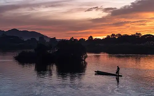 Silhouette d'un pêcheur debout sur sa pirogue au coucher du soleil avec nuages orange, à Don Det.