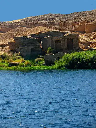 Vue d'un paysage égyptien, de l'eau au premier plan, des grottes et une montagne aride au second plan