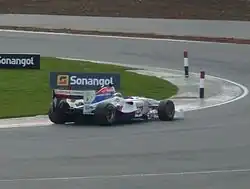 Photographie d'une monoplace blanche, bleue et rouge, vue de trois-quarts dos droit, à l'amorce d'un virage vers la gauche.