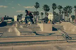 Photo d'un skater en train d'effectuer une figure de skate à Venice Beach. Il est pris en l'air durant un saut à 1 à 2 mètres de hauteur et son skate effectue des rotations en dessous de lui. Le décor est principalement composé d'un multitude d'escaliers en béton de quelques marches. En arrière-plan assez loin, il y a des skaters et beaucoup plus loin, on aperçoit des cocotiers, la plages et des immeubles.