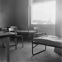 Photo en noir et blanc de l'intérieur d'une chambre. Elles ont des grandes fenêtres et un grand radiateur. Une table et une chaise sont visibles, ainsi que deux lits en métal sur lesquels sont posés des matelas fins.