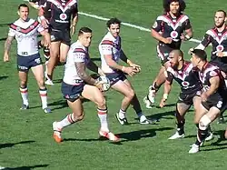 Homme de trois quart, tenant le ballon, avec deux coéquipiers le suivant des yeux. Plusieurs adversaires sont face à lui, sur la droite de la photo.