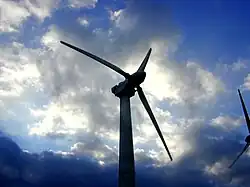 Photographie couleur. Extrémité d'une éolienne avec trois pales se détachant dans un ciel nuageux.