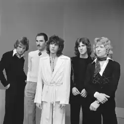 Photo en noir et blanc de cinq jeunes hommes blancs debout côte à côte. Ils ont tous de longs cheveux ondulés ou bouclés, sauf le deuxième, qui a les cheveux courts et une petite moustache ; il jette un regard en coin vers l'objectif.