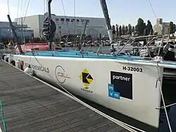 Voilier blanc amarré au ponton, vu de trois quarts avant. Pont bleu, rouf rouge.