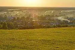 Photographie depuis une colline (le mont Piket) montrant des maisons du village de Strotski, avec de la fumée de cheminée. Le paysage est vert, avec des arbres entre les maisons. La colline est une prairie.