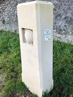 Photographie en couleurs d'une stèle en pierre parallélépipédique avec une coquille Saint-Jacques en bas relief.