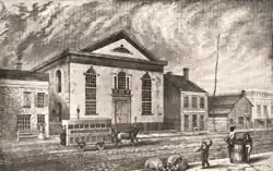 Bâtiment néo-classique. Un tramway hippomobile passe devant le bâtiment, au premier plan, un homme frappe une truie avec une verge pour qu'elle ne fouille pas dans des ordures.