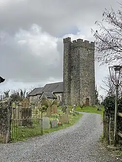 Photo d'un bâtiment allongé dominé par une grande tour carrée à créneaux d'allure massive. Une allée de gravier conduit au bâtiment derrière des rangées de pierres tombales.