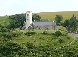 Photo d'un bâtiment allongé aux murs gris et au toit en ardoise doté d'une grande tour carrée blanche sur la gauche, au cœur d'un paysage verdoyant