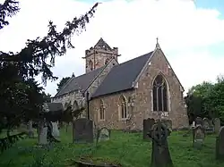 Photo d'un bâtiment aux murs de pierre rouge et au toit en ardoises, avec une tour carrée crénelée, entouré d'une pelouse verte jonchée de pierres tombales