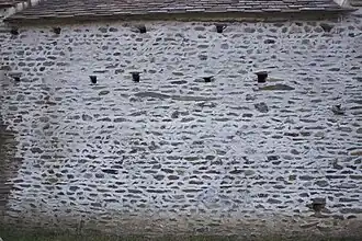 Mur aveugle en pierres percé de petits trous carrés.